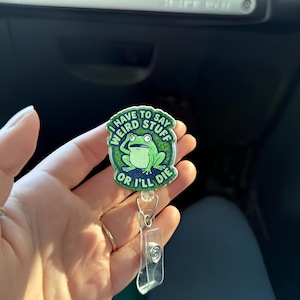 May include: A green and blue badge reel with the text "I HAVE TO SAY WEIRD STUFF OR I'LL DIE" and a cartoon frog. The badge reel has a clear plastic clip and a retractable cord.