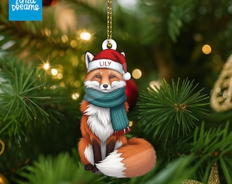 Peut inclure: Ornement de Noël représentant un renard portant un bonnet de Père Noël et une écharpe turquoise. Le renard est orange, blanc et marron, avec le nom "LILY" sur le chapeau. L'ornement est suspendu à une ficelle dorée sur un fond flou de sapin de Noël.