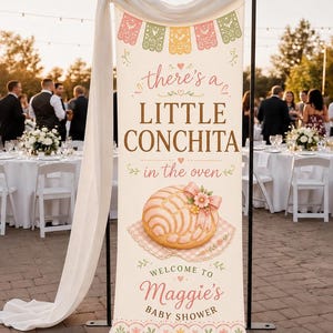 May include: A vertical banner for a baby shower featuring colorful papel picado at the top, the text 'there's a LITTLE CONCHITA in the oven', an illustration of a pink concha pastry with a bow, and 'WELCOME TO Maggie's BABY SHOWER' at the bottom.