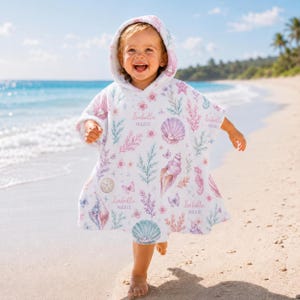 Könnte beinhalten: Ein weißer Kapuzen-Badeponcho mit einem wiederkehrenden Muster aus Aquarell-Muscheln, Korallen und Schmetterlingen in Rosa-, Lila- und Türkistönen. Der Schriftzug Isabella MARIE ist mehrfach in Schreibschrift auf den Stoff gedruckt.