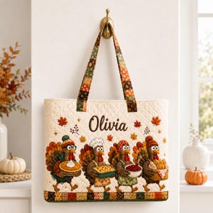 May include: A quilted tote bag featuring the name Olivia in brown script, surrounded by four turkeys wearing hats and carrying Thanksgiving food items, with a patchwork pattern on the handles and bottom edge, hanging from a gold wall hook.