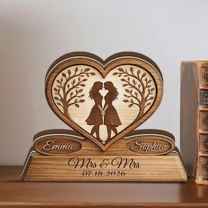 May include: A wooden heart-shaped wedding keepsake with two women silhouettes holding hands, framed by tree designs. The names "Emma" and "Sophia" are engraved, along with "Mrs & Mrs" and the date "07.18.2026".