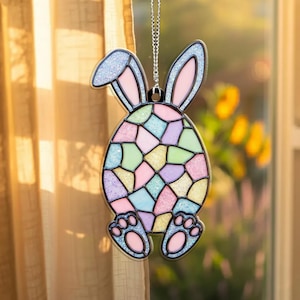 May include: A decorative Easter bunny ornament with a stained glass egg design. The bunny has blue glitter ears and pink inner ears. The egg is composed of pastel-colored geometric shapes. The bunny's feet are at the bottom, and the ornament hangs from a silver chain.