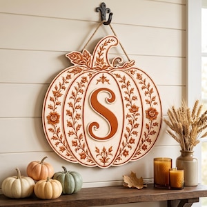 May include: A wall-hanging pumpkin-shaped sign featuring a cream-colored background with embossed orange floral vines and a large orange letter S in the center. The sign is suspended by a rope from a black metal hook mounted on a white wall.