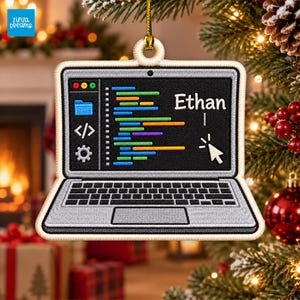 Puede incluir: Un adorno con forma de portátil con un lazo dorado para colgar. La pantalla muestra líneas de código de colores, un icono de carpeta azul, un símbolo de código, un icono de engranaje, un cursor blanco y el nombre Ethan.