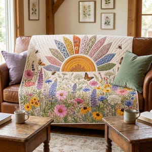 Op de afbeelding: Een crèmekleurige quilt met een groot, veelkleurig zonontwerp met bloembladachtige stralen in diverse bloemmotieven. Het onderste gedeelte toont een levendige weide vol wilde bloemen, waaronder roze, gele en blauwe bloesems, met vlinders en bijen.