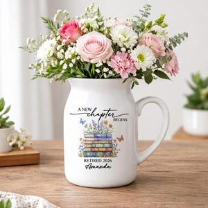 Puede incluir: Jarra de cerámica blanca con asa, llena de un ramo de flores rosas y blancas. La jarra está decorada con el texto "A new chapter begins", una pila de libros, mariposas, el nombre "Amanda" y el año "2026".