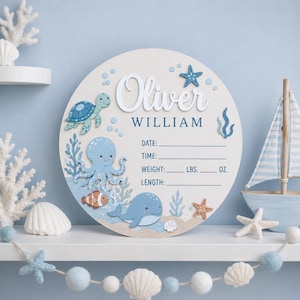Puede incluir: Un letrero circular de madera para nacimiento con el nombre Oliver William en letras blancas y azules. El diseño incluye ilustraciones de una tortuga marina, pulpo, pez payaso y ballena, con líneas para fecha, hora, peso y longitud del bebé.