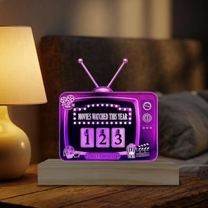 May include: A purple, retro-style TV-shaped cinema log with the text "MOVIES WATCHED THIS YEAR" and the numbers "123" displayed. The base is made of light-colored wood. The log is illuminated, and the text "Chris's Cinema Log" is at the bottom.