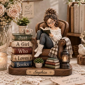 May include: A figurine of a person with curly brown hair and glasses sitting in a brown armchair reading a book. A stack of five books reads Just A Girl Who Loves Books. A nameplate reads Sophia. Includes a lantern, potted plants, and a blanket.