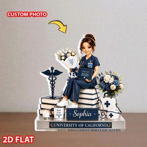 May include: A 2D flat figurine of a nurse in blue scrubs, holding a bouquet and a sign that reads "Class of 2026." The base includes a caduceus, books, a coffee mug, and a medical bag. The text "Sophia" and "University of California" are also present.