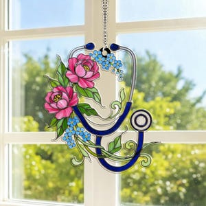 May include: Stained glass stethoscope decoration with pink peonies, blue forget-me-nots, and green leaves. The stethoscope is outlined in blue and silver, with a hanging chain. A decorative piece for medical professionals.