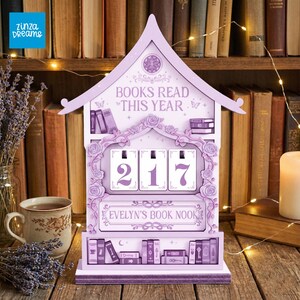 May include: A lavender-colored wooden book counter with the text "BOOKS READ THIS YEAR" and the number "217" displayed. The counter is decorated with floral and book motifs, and the text "EVELYN'S BOOK NOOK" is also visible.