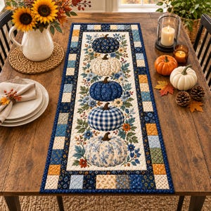 May include: A rectangular quilted table runner featuring a central vertical row of five pumpkins in various blue, white, and cream patterns, surrounded by floral motifs. The border consists of a patchwork of small blue, yellow, and cream squares.