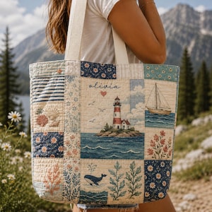 May include: A quilted tote bag featuring a patchwork design with nautical and floral motifs. The center panel displays a lighthouse on an island, a sailboat, a whale, and the name "olivia" with a heart. The fabric includes blue, white, and tan colors.