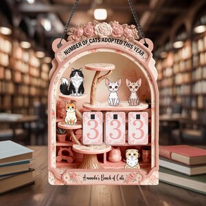 Pode incluir: Uma placa decorativa rosa com tema de gato com o texto "NUMBER OF CATS ADOPTED THIS YEAR" e "Amanda's Bunch of Cats". A placa apresenta ilustrações de gatos e o número "333".