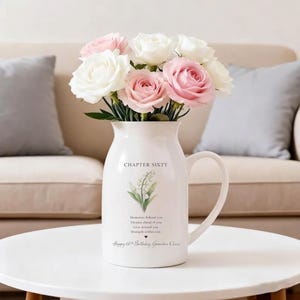 May include: A white ceramic pitcher with a handle, filled with pink and white roses. The pitcher has the text "CHAPTER SIXTY" and a floral design. Below the floral design is the text "Memories behind you, Dreams ahead of you, Love around you, Strength within you, Happy 60th Birthday, Grandma Clara."