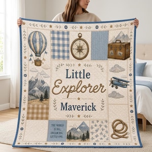 Peut inclure: Une couverture matelassée avec un fond crème et des accents bleus et beiges. Elle présente des illustrations de montgolfière, boussole, valise, avion et corde. Le texte indique Little Explorer Maverick et The world is big, dream big, little one.
