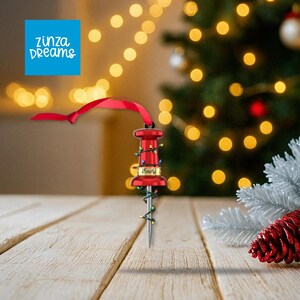 May include: A red pushpin Christmas ornament with the name "Emily" on it. The ornament is decorated with small, colorful lights and a red ribbon for hanging. The background includes a Christmas tree and bokeh lights.