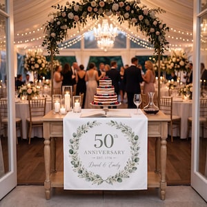 May include: A white table runner featuring a green botanical wreath design with the text 50th ANNIVERSARY EST. 1776 and David & Emily. The runner is displayed on a wooden table with a three-tier berry cake, candles, and an open book in a venue.