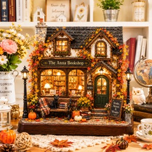 May include: A miniature bookstore model titled The Anna Bookshop, featuring a dark roof, stone chimney, and autumn foliage. The interior is illuminated, showing shelves of books, a black cat on a bench, and a sign reading Books, Stories & Wonder.
