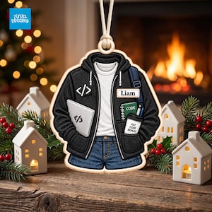 May include: A flat, hanging ornament shaped like a black hoodie over a white shirt and blue jeans. The hoodie features a code bracket icon, a laptop with a code icon, a name tag reading Liam, a green notebook, and a cup labeled Eat Code Repeat.