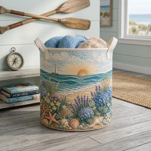 Könnte beinhalten: Ein zylindrischer Aufbewahrungskorb aus Stoff mit einer gesteppten Küstenszene, die einen Sonnenaufgang über dem Meer, ein Segelboot, Muscheln und Lavendel zeigt. Der Korb hat zwei seitliche Griffe und ist mit gefalteten Handtüchern gefüllt.