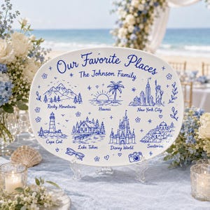 Peut inclure: Un plat ovale en céramique blanche orné d'illustrations au trait bleu représentant des lieux nommés Rocky Mountains, Hawaii, New York City, Cape Cod, Lake Tahoe, Disney World et Santorini. Le texte indique Our Favorite Places et The Johnson Family.