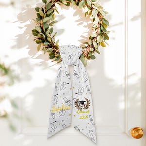 May include: White graduation scarf with black line art of graduation caps and diplomas, accented with gold stars and text. The scarf is personalized with the name "Sophia" and "Class of 2026" in gold lettering. A green eucalyptus wreath hangs in the background.