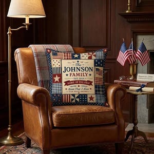 May include: A brown leather armchair with a decorative pillow featuring a patriotic design. The pillow has a patchwork pattern with stars and stripes, and the text "The JOHNSON FAMILY EST. 2024 HOME OF THE FREE" is displayed. A plaid blanket is draped over the chair.