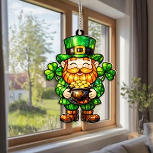 May include: A St. Patrick's Day window decoration depicting a leprechaun with a pot of gold. The leprechaun is dressed in green attire, including a hat, jacket, and trousers, and has a red beard and shamrocks. It hangs from a silver chain.