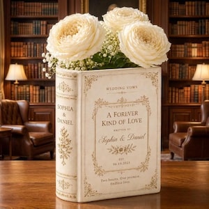 May include: A cream-coloured book-shaped vase featuring gold-toned decorative borders and text that reads: WEDDING VOWS, A FOREVER KIND OF LOVE, WRITTEN BY Sophia & Daniel, EST 06.14.2025, Two hearts. One promise. Endless love. Filled with white roses.