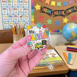 Può includere: Un portabadge circolare con un design scolastico colorato e il testo Ms. OLIVIA. La grafica include uno scuolabus giallo, una pila di libri, un mappamondo, un arcobaleno, una stella e una piccola lavagna con il testo Teach Love Inspire.