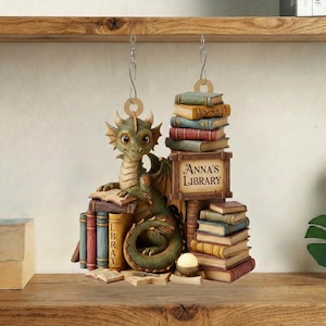 May include: A decorative dragon ornament featuring a stack of books and a sign reading "Anna's Library." The dragon is green and brown, with a friendly face. The books are in various colours, and the ornament hangs from a chain.