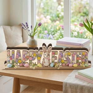 May include: A wooden "Happy Easter" sign with a bunny and floral accents. The sign features the words "Happy Easter" in a light-up design, with a bunny figurine and colorful eggs. The background includes a window with flowers and a table with books and vases.