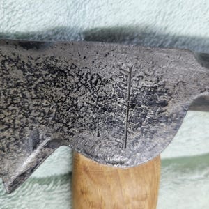 Op de afbeelding: Close-up van een vintage bijlkop met een houten handvat. De metalen kop heeft een getextureerd, verweerd uiterlijk met zichtbare markeringen. De houten handgreep is lichtbruin.