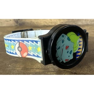 Könnte beinhalten: Eine schwarz-weiße Pokemon-Uhr mit einer Bulbasaur-Grafik auf dem Zifferblatt. Das Uhrenarmband ist weiß mit blauen Zickzacklinien, einem rot-weißen Pokeball und gelben Sternen. Die Uhr liegt auf einer braunen Oberfläche.
