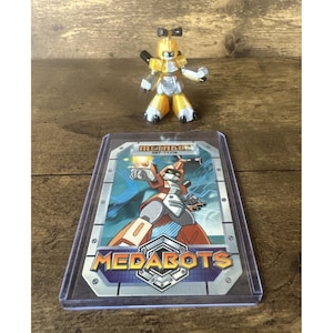 May include: A yellow and silver Medabot action figure stands above a Medabots trading card. The card features a robot illustration with the word "MEDABOTS" prominently displayed. The action figure has articulated limbs.
