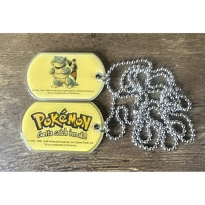 May include: Two yellow Pokémon dog tags with a silver ball chain. One tag features a cartoon turtle-like character. The other tag has the Pokémon logo and the phrase "Gotta catch 'em all!" The tags are made of a shiny material.