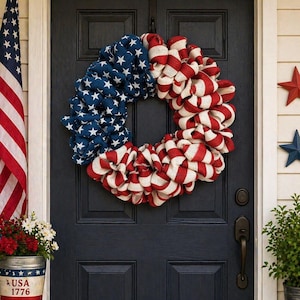 May include: A patriotic wreath with a blue section featuring white stars and a red and white striped section, hanging on a black door. An American flag and a bucket of flowers are visible.