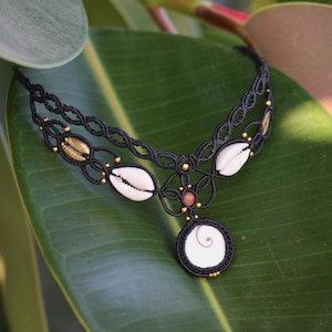 Puede incluir: Collar de macramé negro con conchas blancas, detalles dorados y un colgante circular con un diseño en espiral. El collar se muestra sobre una gran hoja verde, resaltando su estilo natural y bohemio.