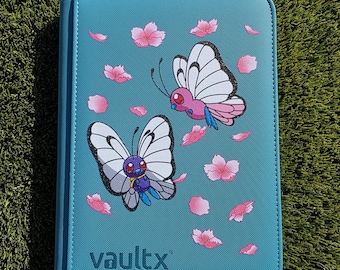 May include: Teal card binder with a zipper closure, featuring illustrations of two cartoon butterflies and pink cherry blossom petals. The text "vaultx" is printed on the lower right corner.
