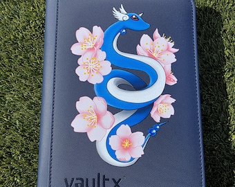 May include: A navy blue card binder with a stylized blue and white serpent design intertwined with pink cherry blossoms. The binder has the word "vaultx" printed in white at the bottom.