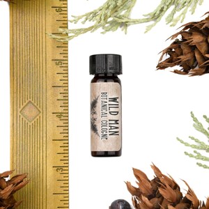 May include: A small amber glass bottle of Wild Man Botanical Cologne with a black cap, positioned next to a yellow ruler showing measurements in inches. The bottle features a tan label with black text and a pine tree graphic illustration.