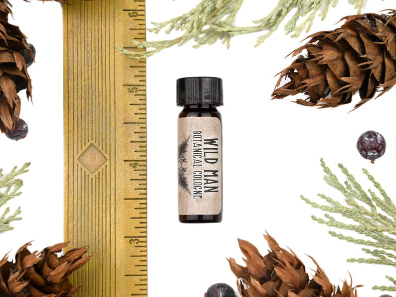 May include: A small amber glass bottle of Wild Man Botanical Cologne with a black cap, positioned next to a yellow ruler showing measurements in inches. The bottle features a tan label with black text and a pine tree graphic illustration.