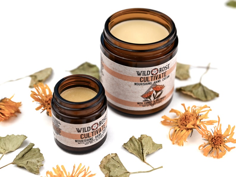 Cultivate Nourishing Hand Cream in amber glass jars. Caps are removed revealing a creamy light orange balm. Dried calendula and gotu kola surround.