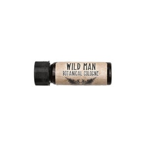 May include: A small amber glass bottle of cologne with a black screw-top cap. The label is light brown with a textured appearance and features the black text WILD MAN BOTANICAL COLOGNE above a small graphic illustration of two pine branches.