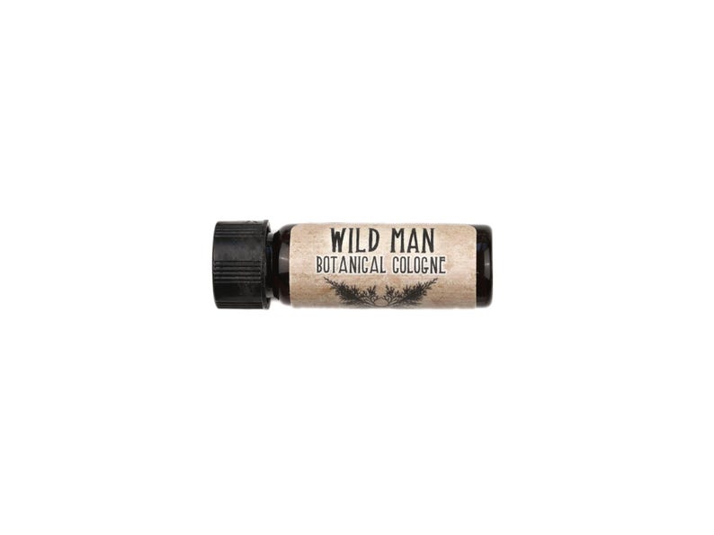 May include: A small amber glass bottle of cologne with a black screw-top cap. The label is light brown with a textured appearance and features the black text WILD MAN BOTANICAL COLOGNE above a small graphic illustration of two pine branches.