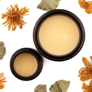 Cultivate Nourishing Hand Cream in amber glass jars. Caps are removed revealing a creamy light orange balm. Dried calendula and gotu kola surround.