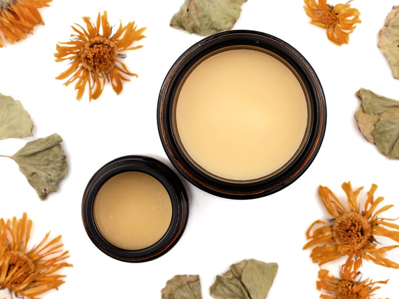 Cultivate Nourishing Hand Cream in amber glass jars. Caps are removed revealing a creamy light orange balm. Dried calendula and gotu kola surround.